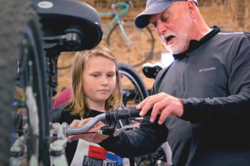 An older man guides a young person as they examine bicycle handlebars in a bike repair workshop, with additional bikes visible in the background.