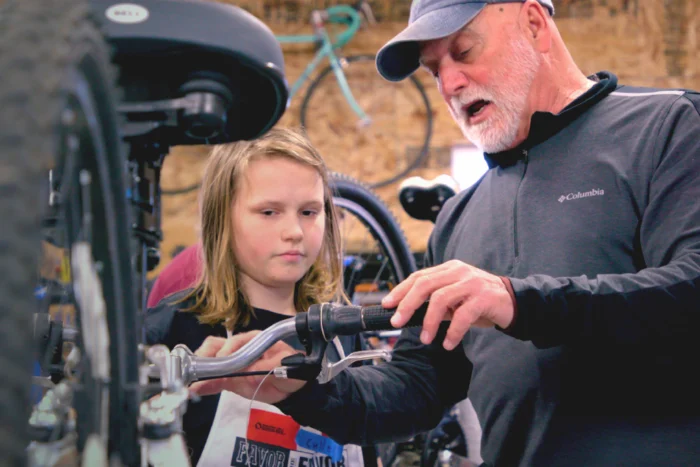 An older man guides a young person as they examine bicycle handlebars in a bike repair workshop, with additional bikes visible in the background.