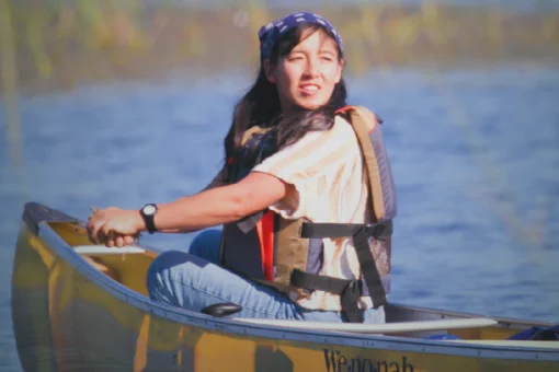 A woman wearing a life vest and bandana paddling a yellow Wenonah canoe on a calm lake surrounded by autumn wetland grasses.