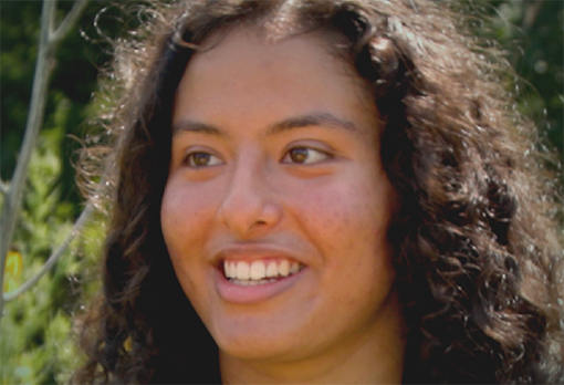 Headshot of Aishika, smiling warmly with curly dark hair, photographed outdoors with green foliage in the background.