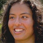 Headshot of Aishika, smiling warmly with curly dark hair, photographed outdoors with green foliage in the background.