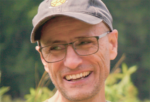 Headshot of Claudio Gratton, smiling warmly, wearing glasses and a baseball cap, photographed outdoors with green foliage in the background.