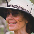 Headshot of Deirdre, smiling, wearing a wide-brimmed sun hat and sunglasses, photographed outdoors.