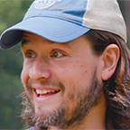 Headshot of James Crall, a smiling man with long brown hair and a beard wearing a blue baseball cap, with trees in the background.