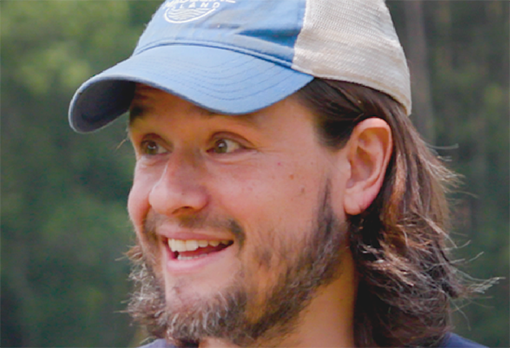 Headshot of James Crall, a smiling man with long brown hair and a beard wearing a blue baseball cap, with trees in the background.