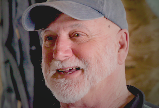 Joe Horvath smiles warmly during an interview, wearing a gray baseball cap against a softly lit workshop background.