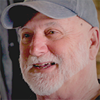 Joe Horvath smiles warmly during an interview, wearing a gray baseball cap against a softly lit workshop background.