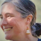 Portrait of Gretchen Gerrish smiling in profile, with gray-streaked hair pulled back and handcrafted earrings, set against a soft green outdoor background.