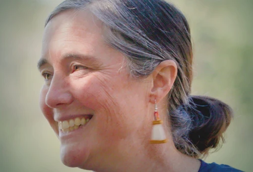 Portrait of Gretchen Gerrish smiling in profile, with gray-streaked hair pulled back and handcrafted earrings, set against a soft green outdoor background.