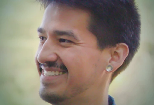 Portrait of Ray Allen smiling in profile, with short dark hair and a small gauge earring, set against a soft green outdoor background.