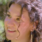 Portrait of Sagen Quale smiling with curly hair and a delicate nose ring, wearing long earrings, set against a blurred green outdoor background.