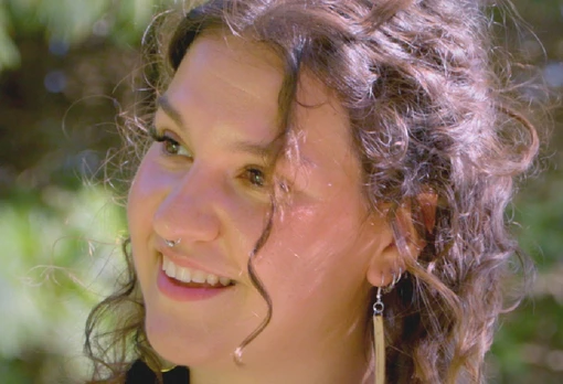 Portrait of Sagen Quale smiling with curly hair and a delicate nose ring, wearing long earrings, set against a blurred green outdoor background.