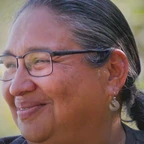 Portrait of Kathy Smith smiling in profile, with long dark hair pulled back, wearing glasses and small hoop earrings, set against a soft green outdoor background.