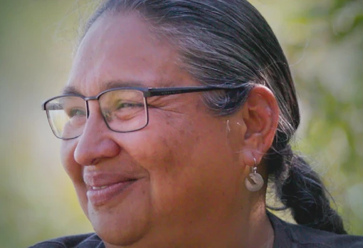Portrait of Kathy Smith smiling in profile, with long dark hair pulled back, wearing glasses and small hoop earrings, set against a soft green outdoor background.