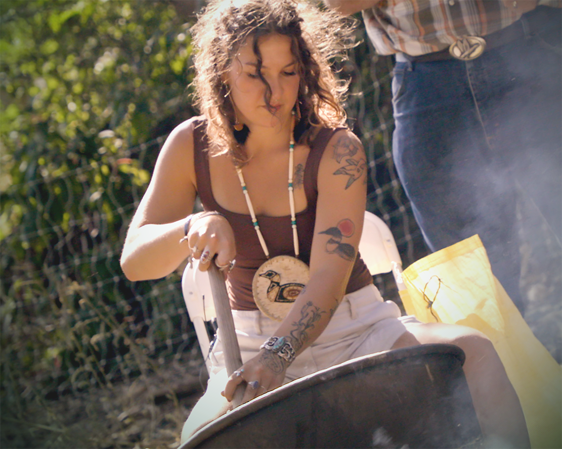A woman with curly hair and tattoos leans over a large cast iron pot outdoors, wearing a traditional bird pendant necklace, with smoke rising around her.