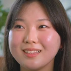 Portrait of Esther Ko smiling warmly, with straight dark hair, set against a soft blurred indoor background with greenery.