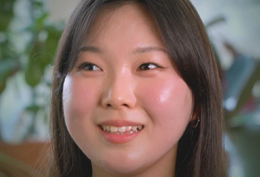 Portrait of Esther Ko smiling warmly, with straight dark hair, set against a soft blurred indoor background with greenery.
