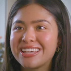 Portrait of Reya Khatri smiling brightly, with long straight dark hair and small hoop earrings, set against a soft indoor background with plants.