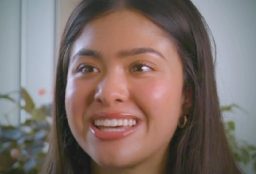 Portrait of Reya Khatri smiling brightly, with long straight dark hair and small hoop earrings, set against a soft indoor background with plants.