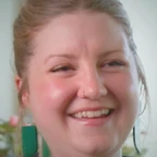 Portrait of Taylor Seale laughing, with blond hair pulled back, a nose ring, and green rectangular earrings, set against a light background with soft floral details.