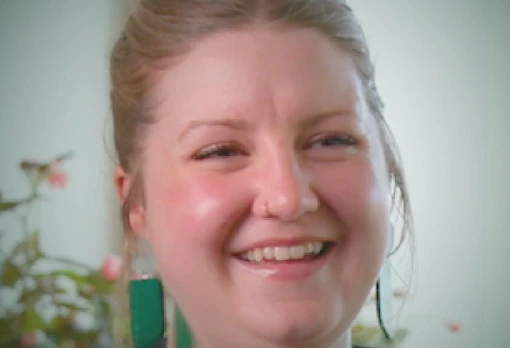 Portrait of Taylor Seale laughing, with blond hair pulled back, a nose ring, and green rectangular earrings, set against a light background with soft floral details.