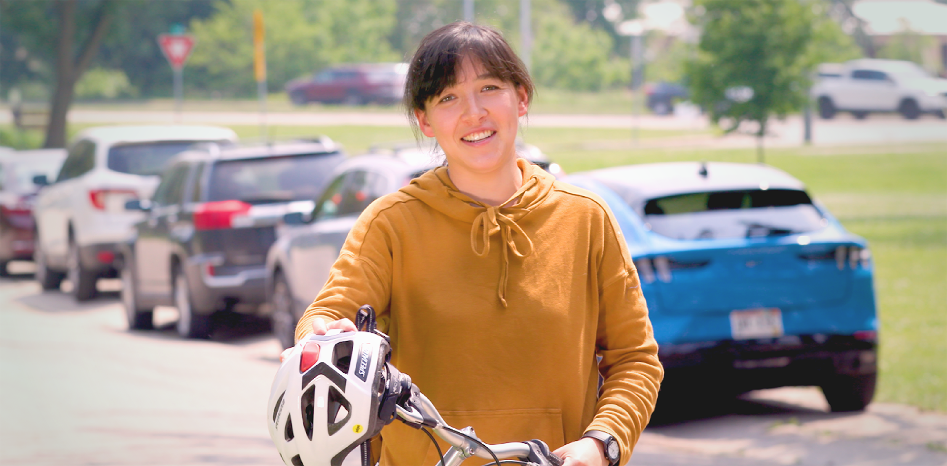A smiling woman in a mustard hoodie holds a bicycle and helmet on a residential street, with parked cars visible behind her.