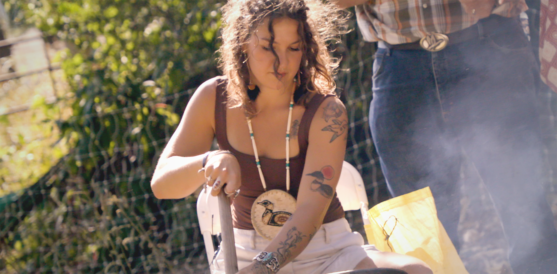 A woman with curly hair and tattoos leans over a large cast iron pot outdoors, wearing a traditional bird pendant necklace, with smoke rising around her.