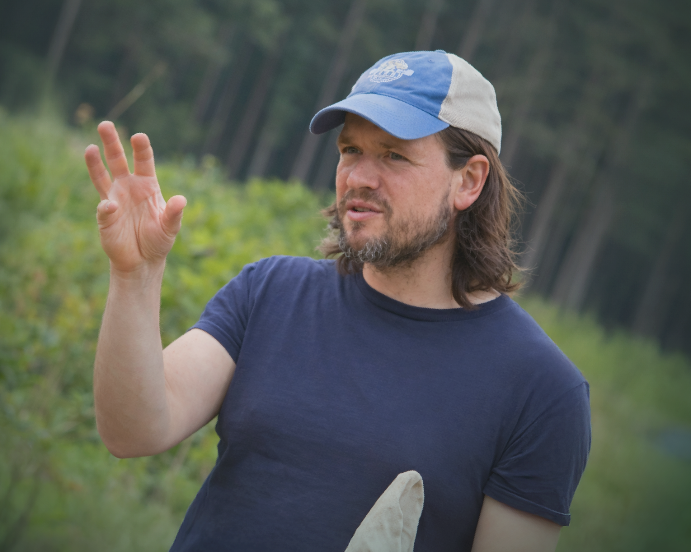 A man with shoulder-length brown hair and a beard, wearing a blue baseball cap and navy t-shirt, gestures expressively with one hand while speaking outdoors, with lush green vegetation in the background.