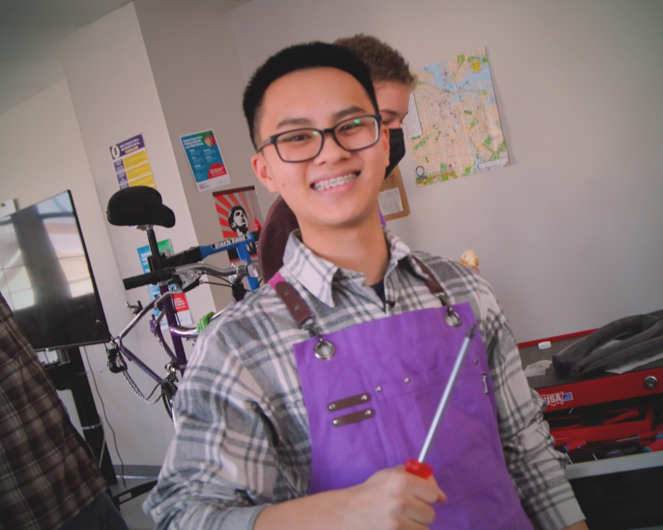 A young person with glasses and braces smiles while holding a screwdriver. They wear a plaid shirt and purple apron in a workshop with a bike and map.