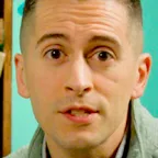 Person in green quilted pullover speaking to camera with bookshelf and teal wall behind