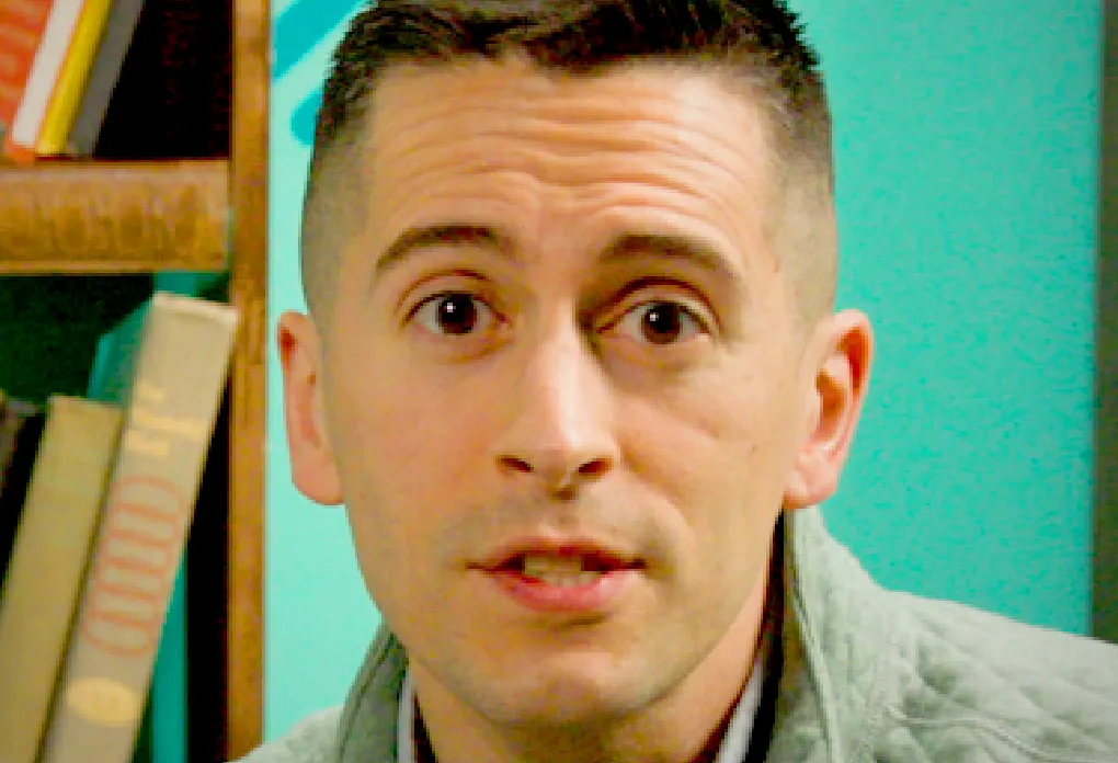 Person in green quilted pullover speaking to camera with bookshelf and teal wall behind