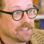 Person with round glasses, mustache, and checkered blazer speaking in a colorful bookstore