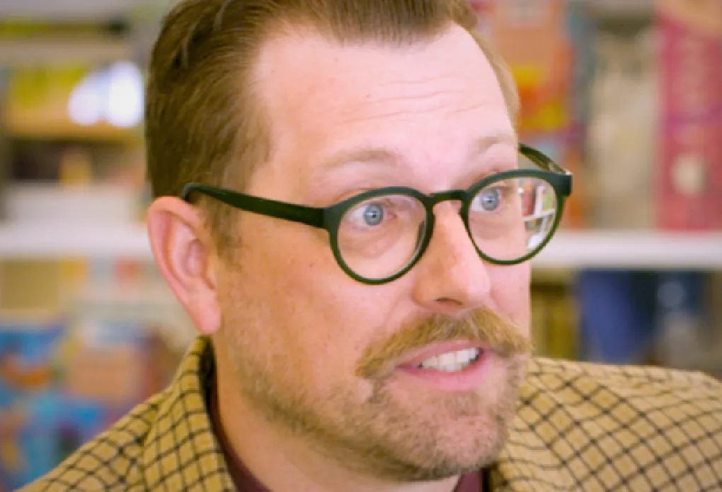 Person with round glasses, mustache, and checkered blazer speaking in a colorful bookstore