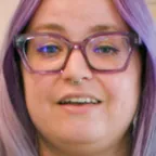 Person with purple hair, glasses, and nose ring smiling at camera with framed art behind