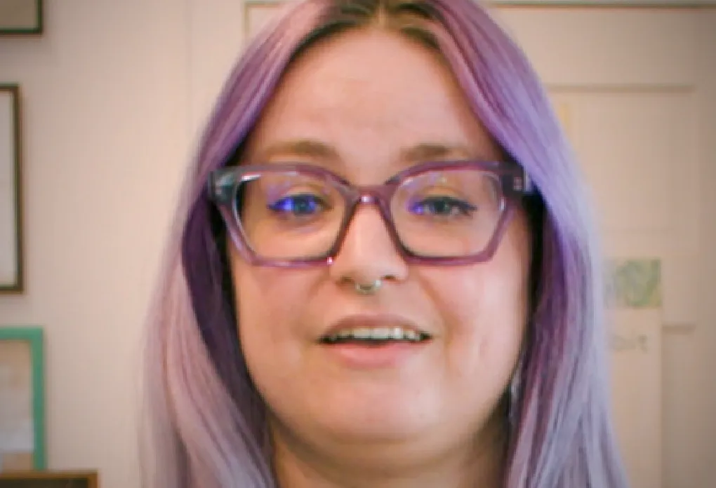 Person with purple hair, glasses, and nose ring smiling at camera with framed art behind