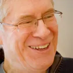 Person with white hair and glasses laughing while looking down, wearing navy zip pullover