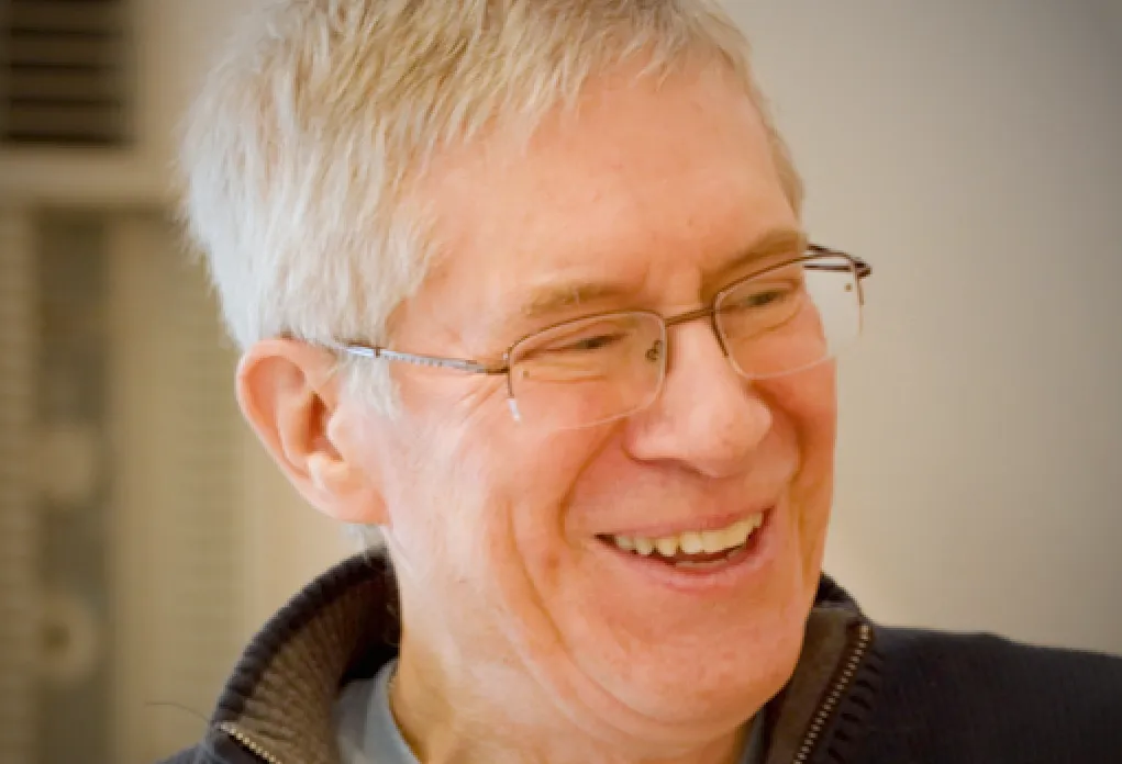 Person with white hair and glasses laughing while looking down, wearing navy zip pullover