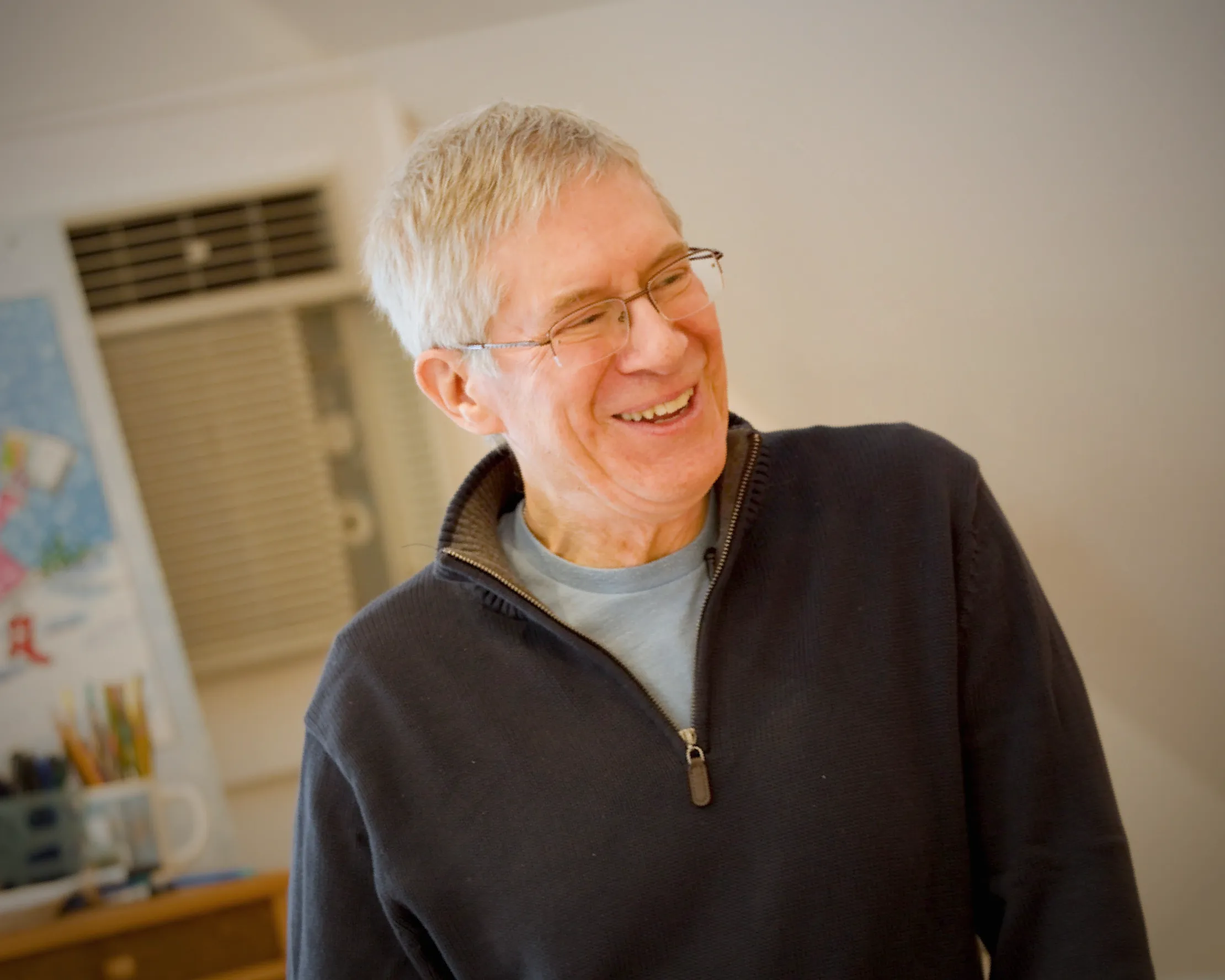 Person with white hair and glasses laughing in a studio with art supplies on a desk behind