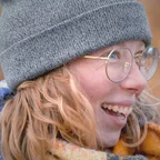 A young woman with blonde-red hair, round gold-framed glasses, and a gray knit beanie laughs while looking to the side, wearing a colorful scarf outdoors.