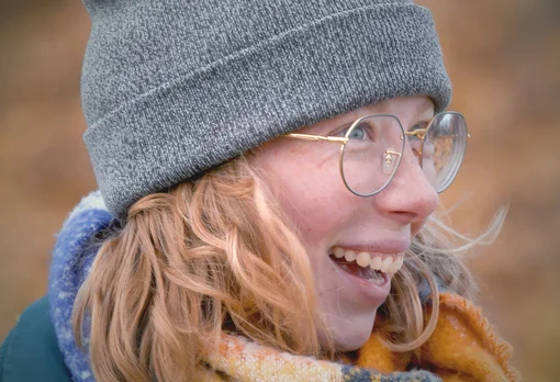 A young woman with blonde-red hair, round gold-framed glasses, and a gray knit beanie laughs while looking to the side, wearing a colorful scarf outdoors.