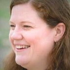 A woman with long auburn hair smiles warmly while looking to the side, photographed outdoors against a soft green background.