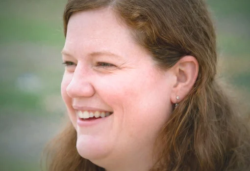 A woman with long auburn hair smiles warmly while looking to the side, photographed outdoors against a soft green background.
