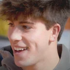 A young man with short dark hair and a gray hoodie smiles while looking to the side, photographed outdoors with a blurred colorful background.