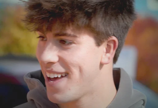 A young man with short dark hair and a gray hoodie smiles while looking to the side, photographed outdoors with a blurred colorful background.
