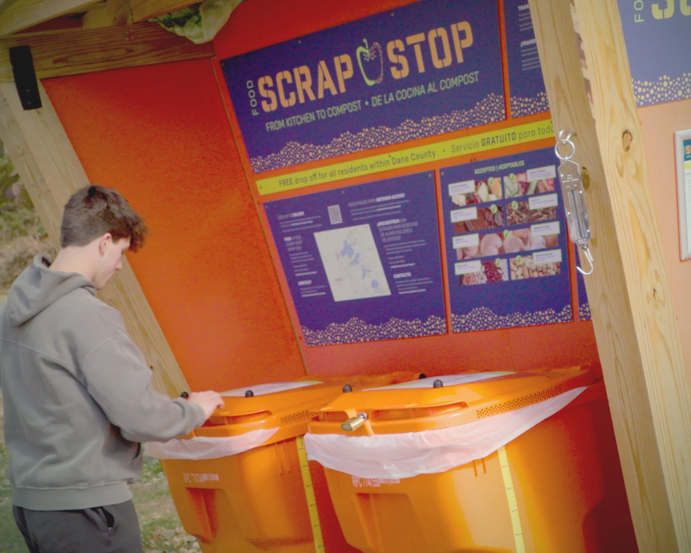 A teenager in a gray hoodie deposits food scraps into a bright orange bin at a 