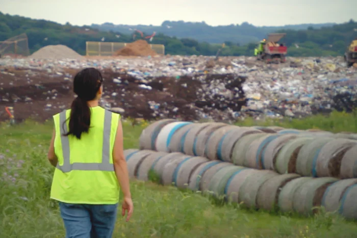 A worker in a yellow high-visibility vest walks along a grassy embankment beside large corrugated pipes, overlooking an active landfill with heavy machinery operating in the background.