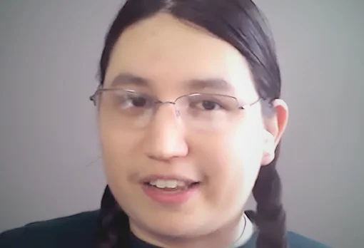 Headshot of guest Daniel Hayden. They have long dark hair in two braids and wire-rimmed glasses, and are speaking during a video call against a neutral grey background.