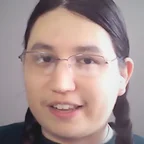 Headshot of guest Daniel Hayden. They have long dark hair in two braids and wire-rimmed glasses, and are speaking during a video call against a neutral grey background.