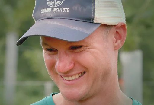 Headshot of guest Nate Lawrence. They are smiling and looking slightly downward, wearing a grey and white Savanna Institute trucker cap and a teal shirt, photographed outdoors with a soft green background.