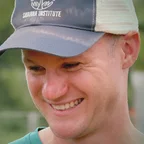 Headshot of guest Nate Lawrence. They are smiling and looking slightly downward, wearing a grey and white Savanna Institute trucker cap and a teal shirt, photographed outdoors with a soft green background.
